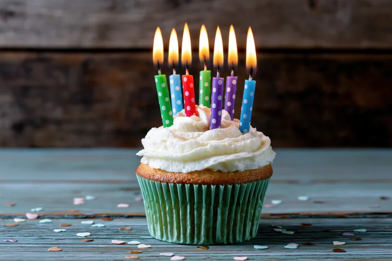Single birthday cupcake with lit colorful candles and white frosting on wooden table
