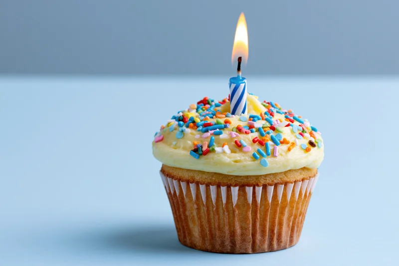 Single lit birthday candle on a frosted cupcake with colorful sprinkles on a blue surface