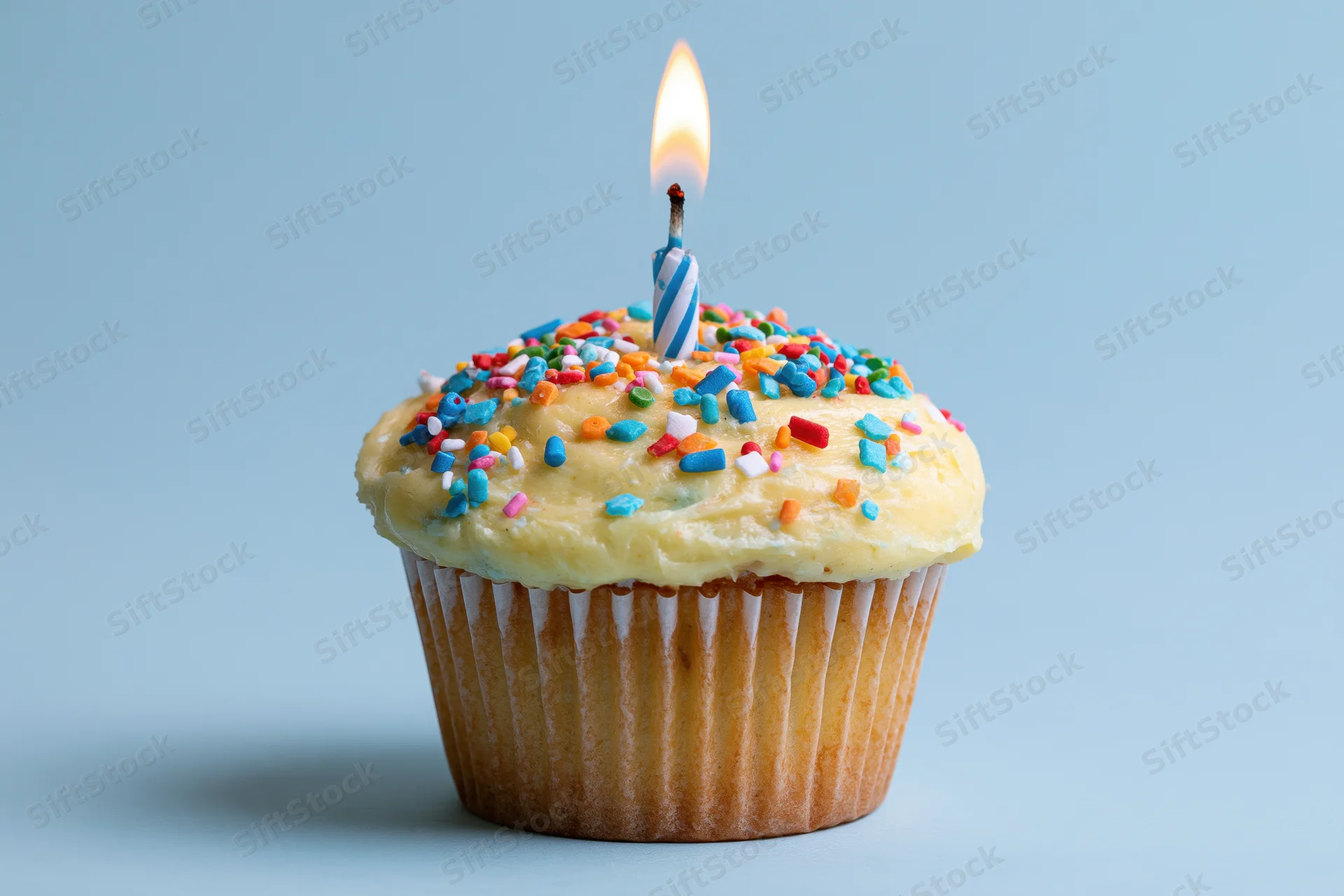 Single cupcake with lit blue-white striped candle and colorful sprinkles on light blue background