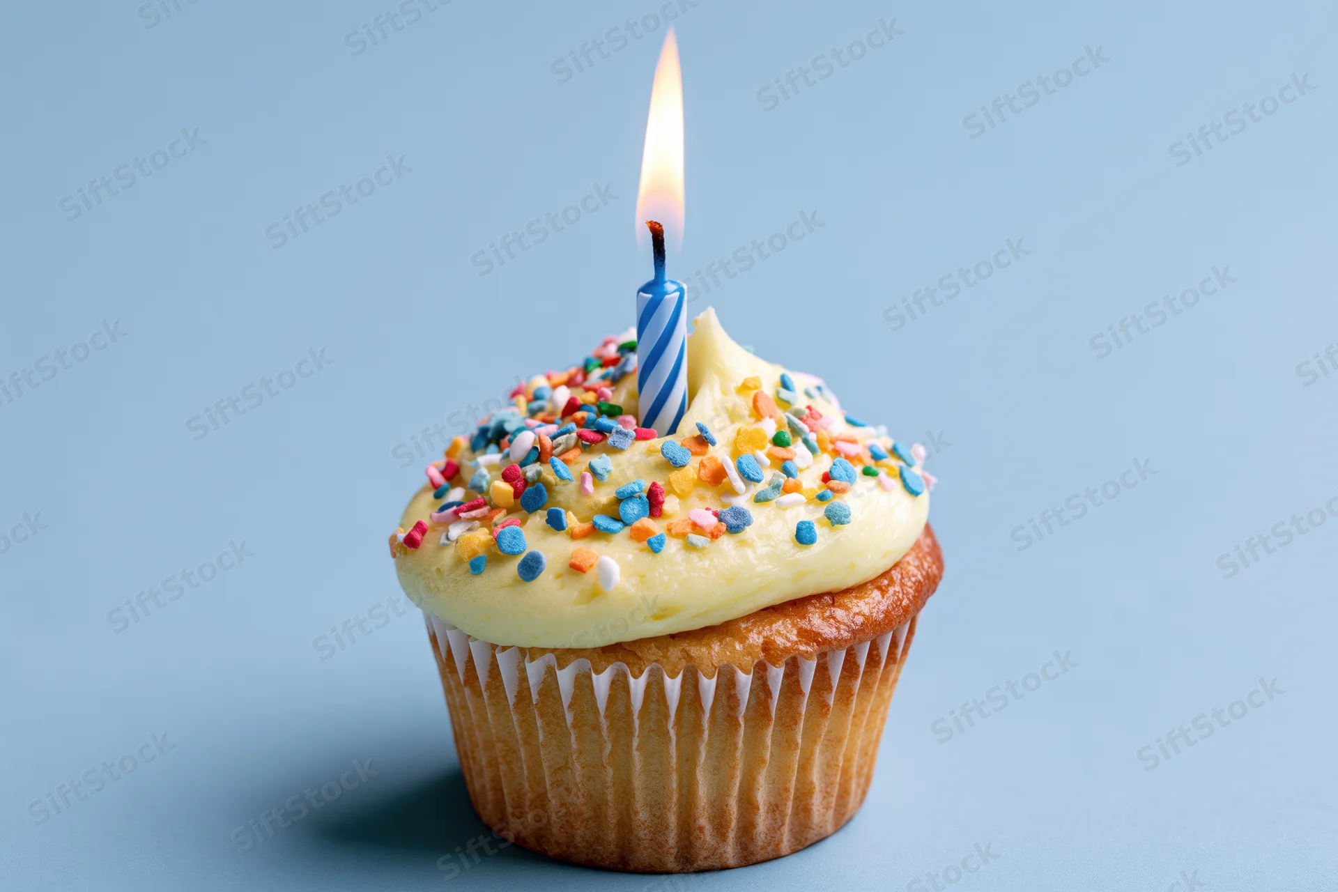 A cupcake with yellow frosting and colorful sprinkles topped with a lit blue and white spiral candle on a plain blue background