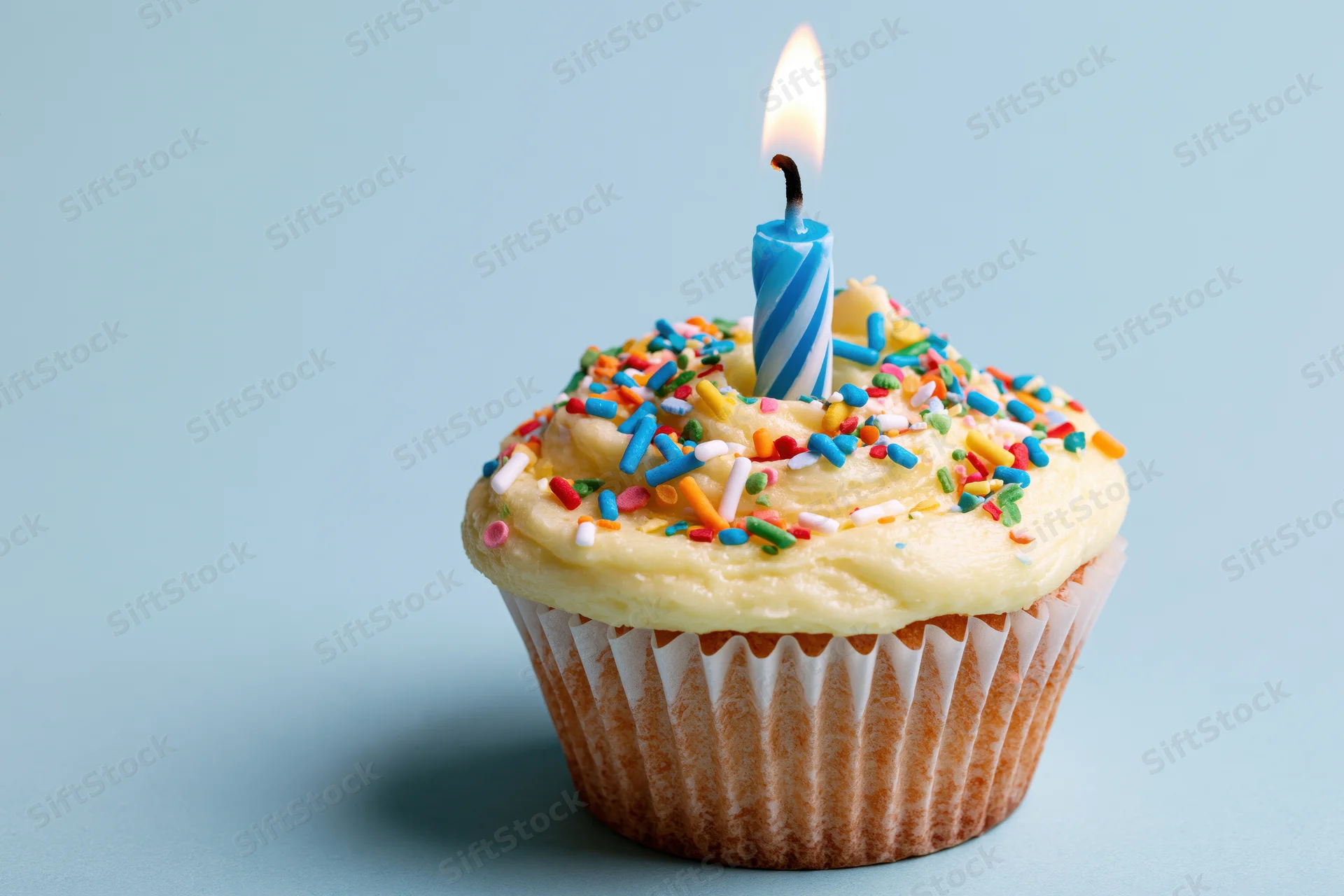 A cupcake with yellow frosting and colorful sprinkles topped with a lit blue and white spiral candle on a light blue background