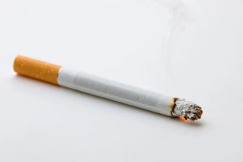 Lit cigarette with white smoke and ash resting horizontally on white surface