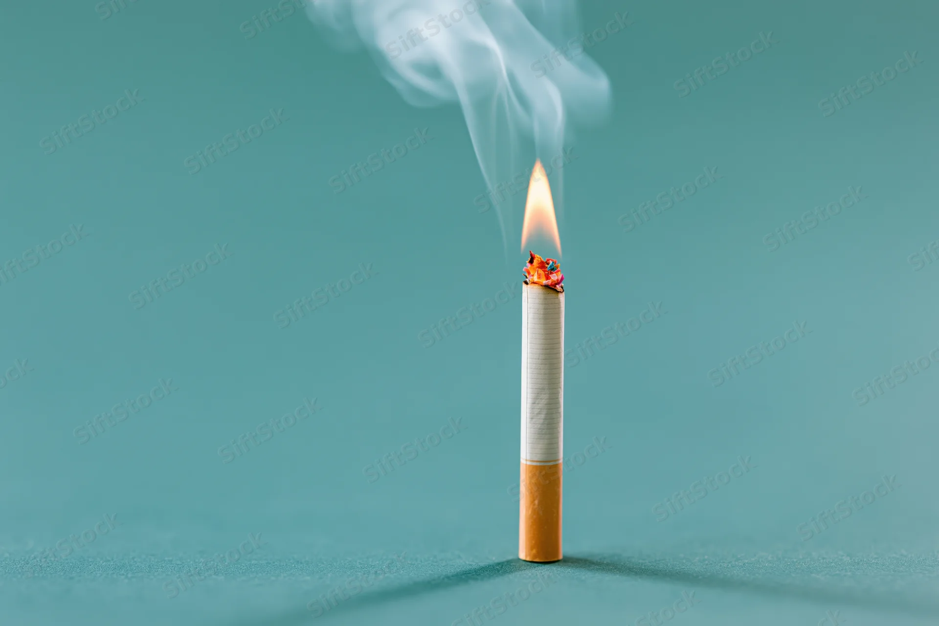 Lit cigarette burning upright on a flat teal surface with smoke rising
