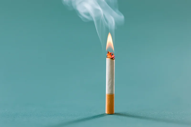 Lit cigarette burning upright on a flat teal surface with smoke rising