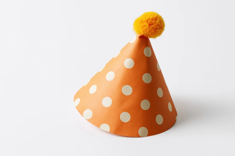Orange paper party hat with white polka dots and yellow pompon on white background