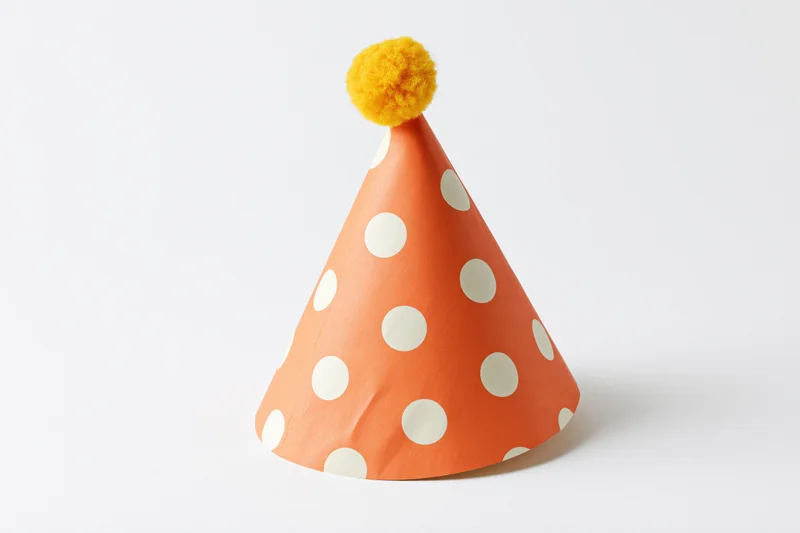 Orange paper party hat with white polka dots and yellow pompon on white background