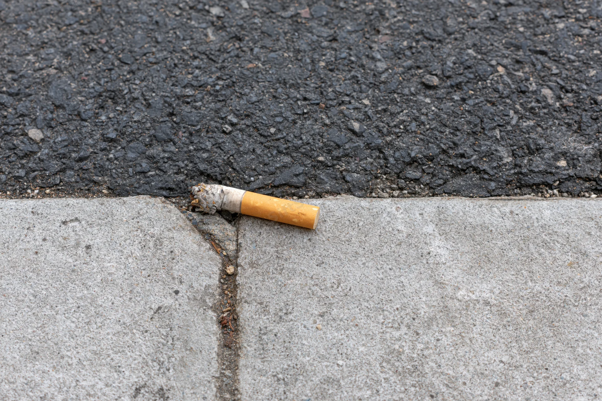 Used cigarette butt resting on the edge between asphalt and concrete curb