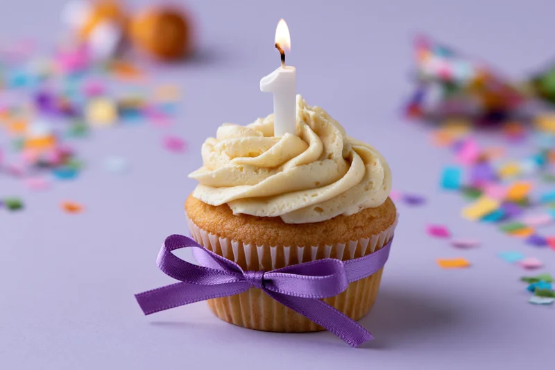 Vanilla cupcake with swirled cream frosting and lit number one candle, tied with purple ribbon, surrounded by colorful confetti on lavender background