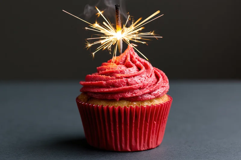 Single vanilla cupcake with swirled red frosting and lit sparkler on dark surface