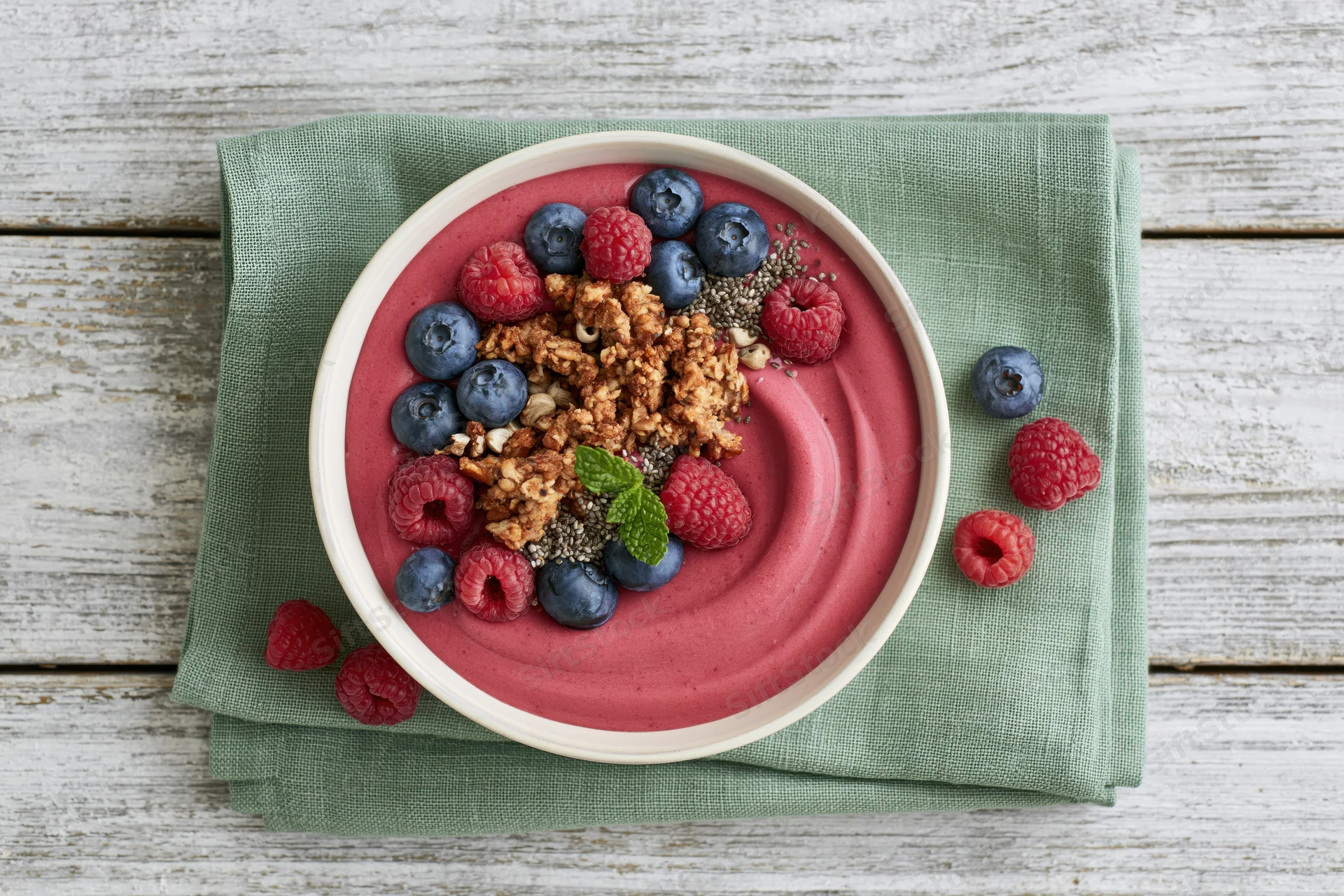 Smoothie bowl with pink berry puree topped with granola, blueberries, raspberries and mint on a green napkin over a wooden table