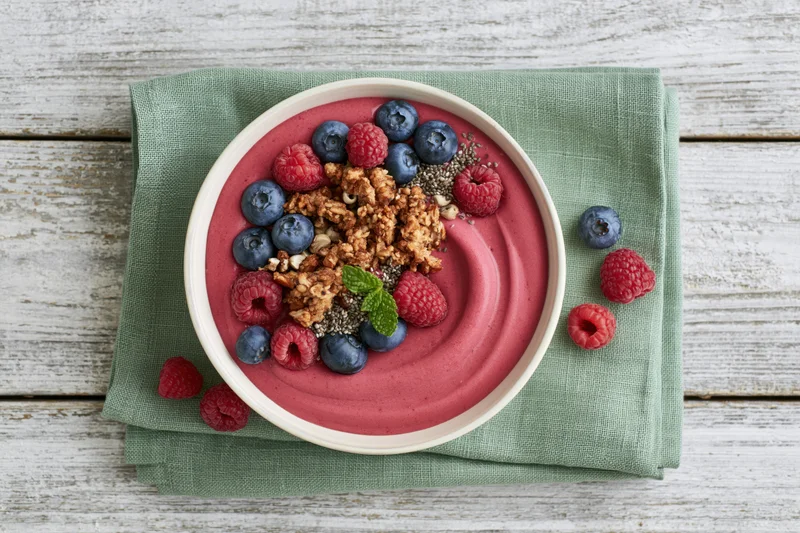 Smoothie bowl with pink berry puree topped with granola, blueberries, raspberries and mint on a green napkin over a wooden table