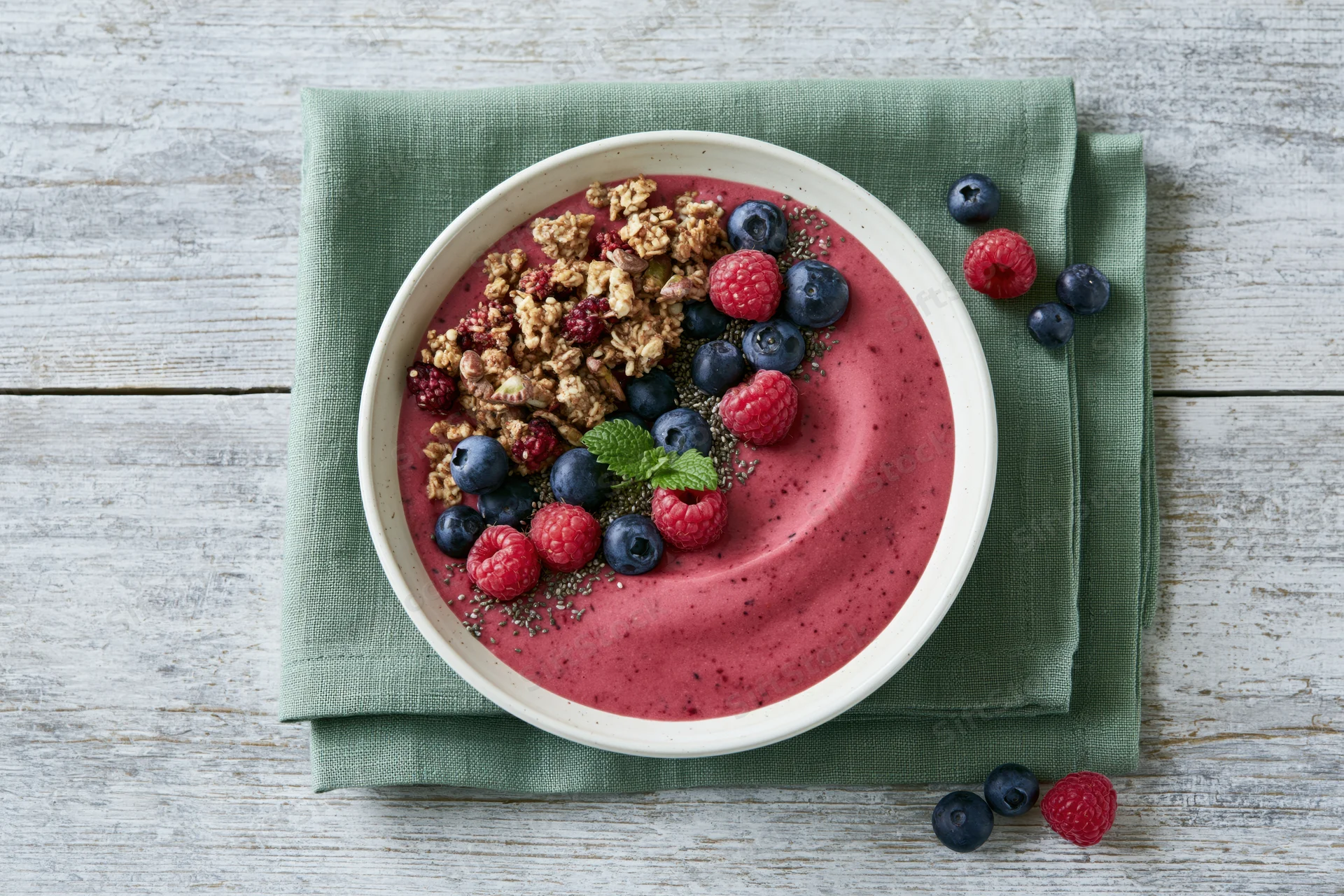 Smoothie bowl topped with granola, berries and mint on a green napkin over a wooden table