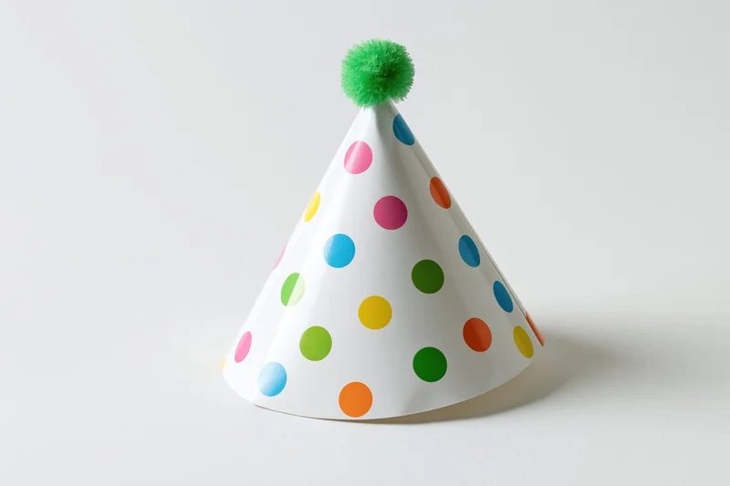 White paper party hat with multicolored polka dots and green pompon on plain surface