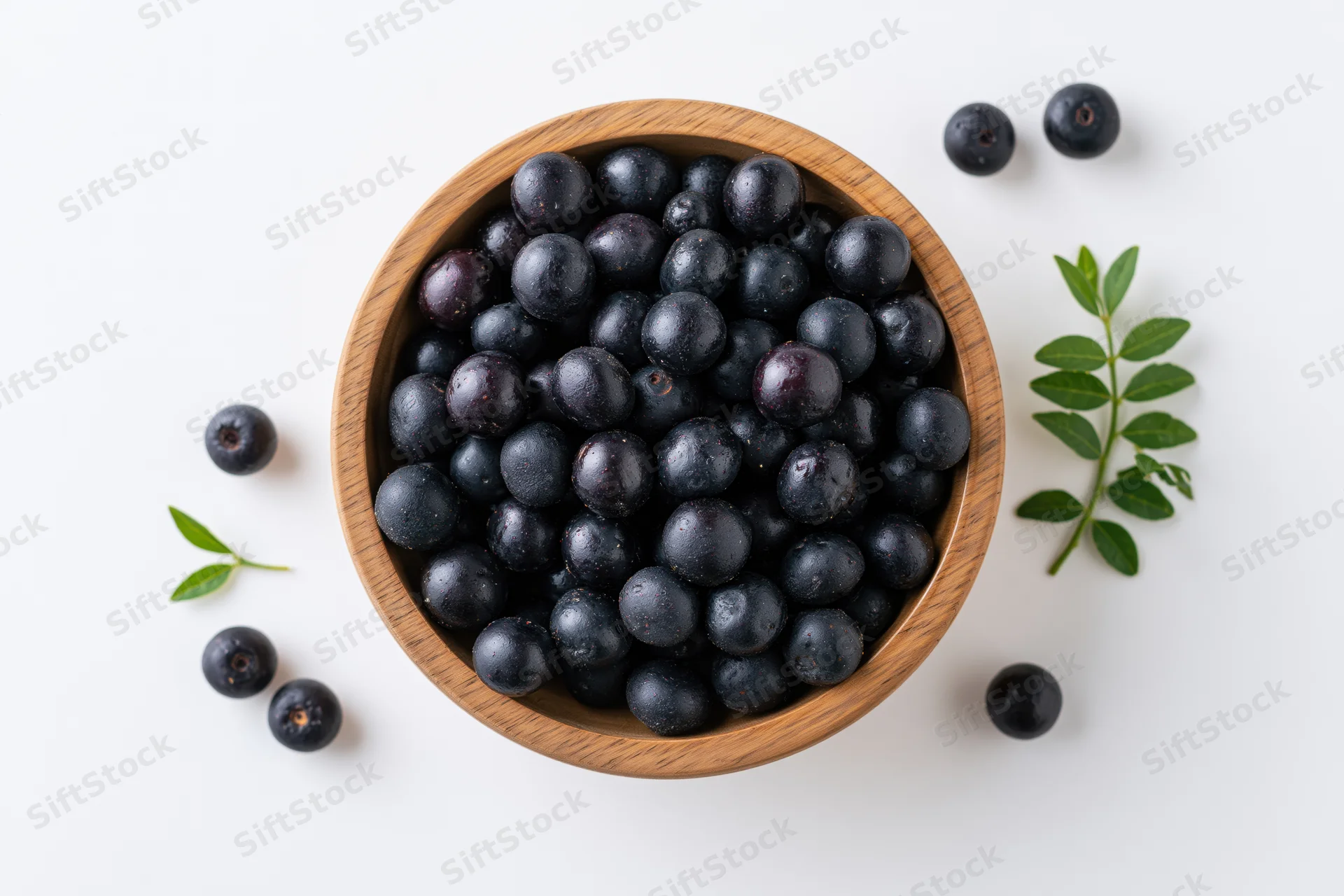 Acai berries in a wooden bowl with scattered berries and green leaves on white background