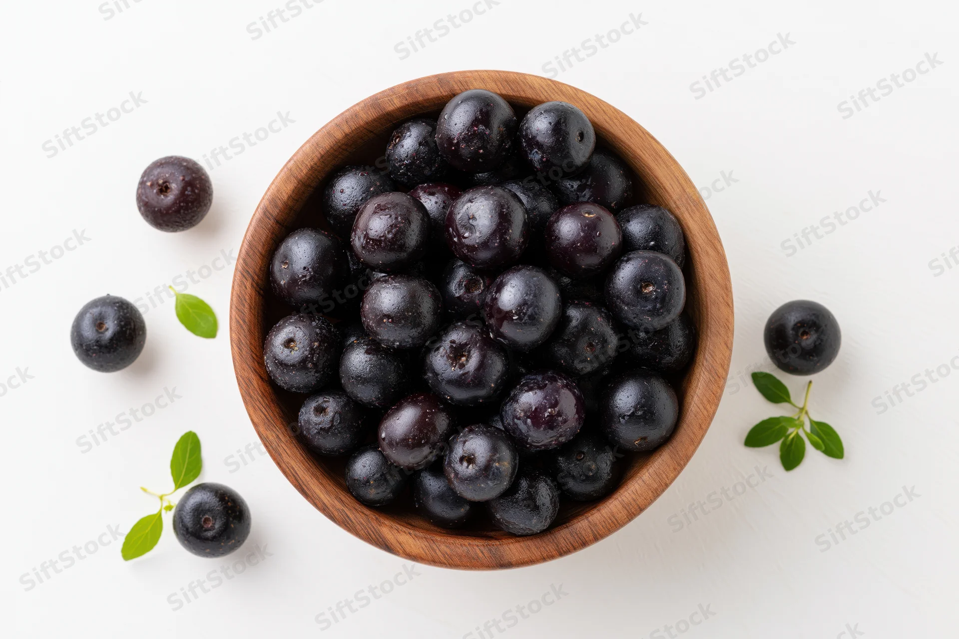 Acai berries in a wooden bowl with scattered berries and green leaves on white surface