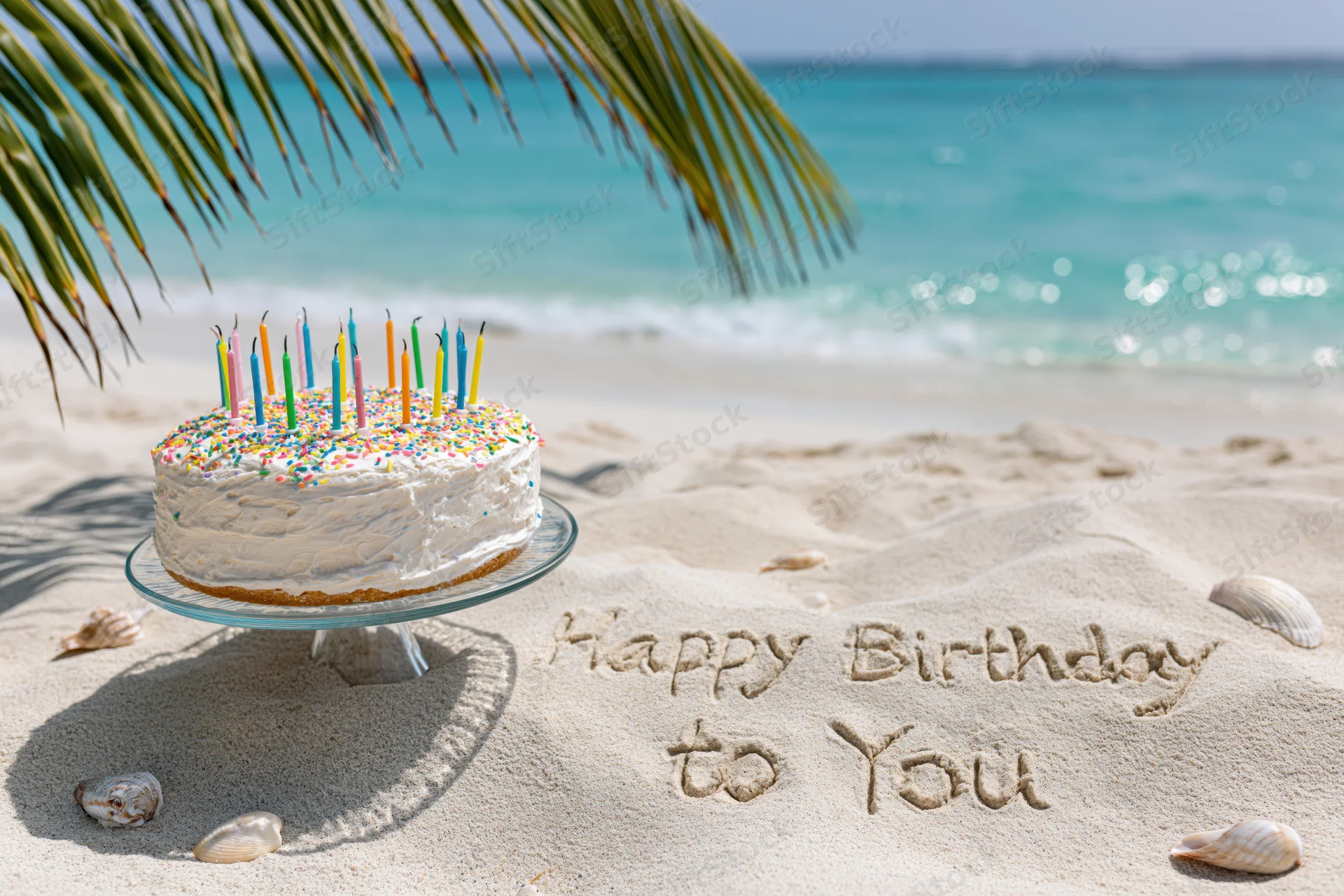 Birthday cake with colorful candles on glass stand on sandy beach near ocean, with happy birthday written in sand and palm fronds overhead