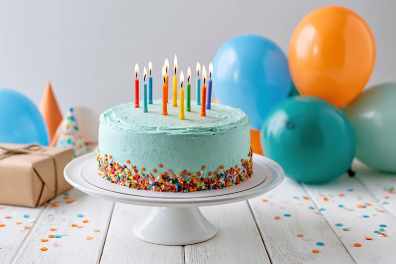 Birthday cake with lit colorful candles on a white cake stand surrounded by balloons and gifts