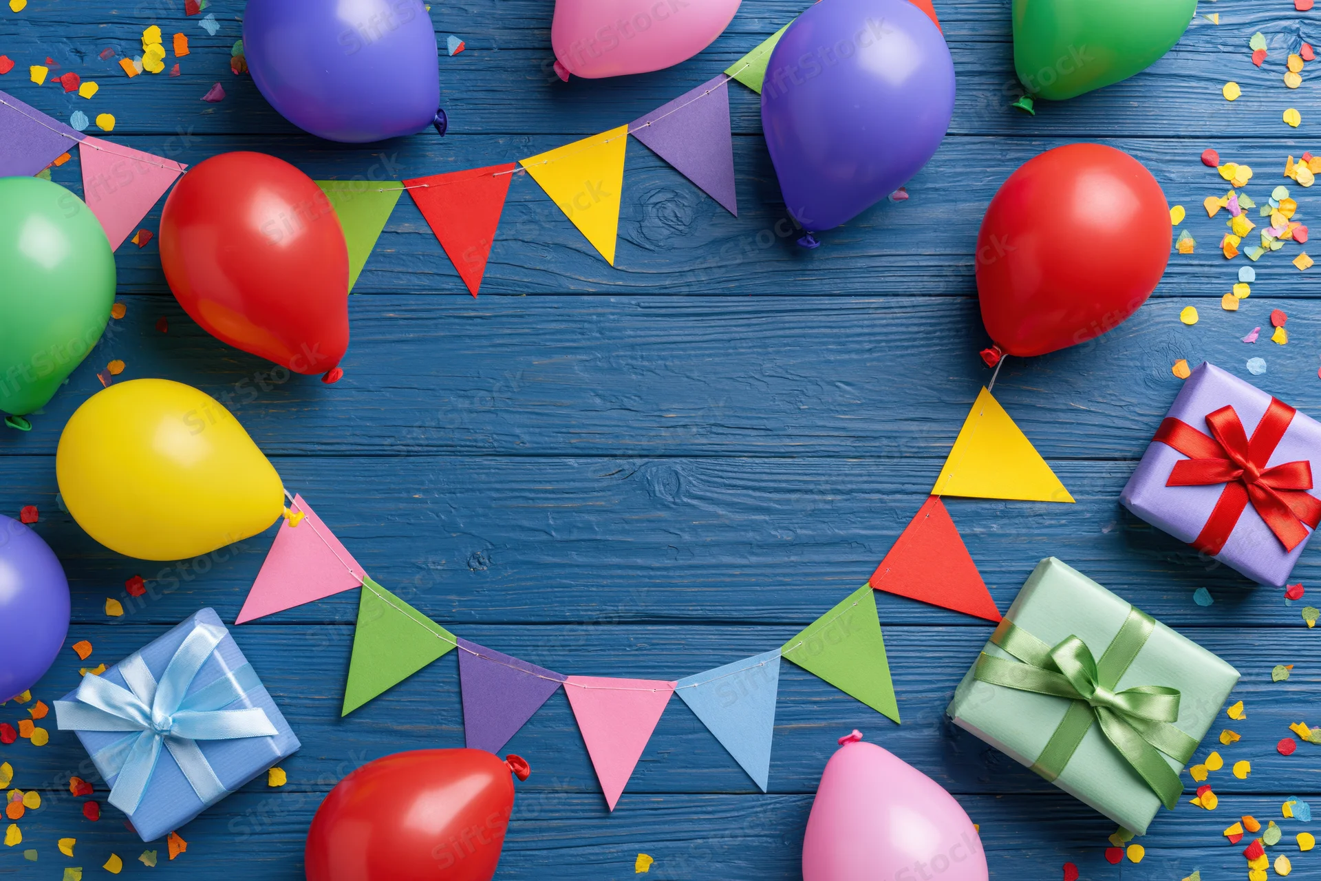 Colorful balloons and wrapped gifts arranged with bunting and confetti on blue wooden surface