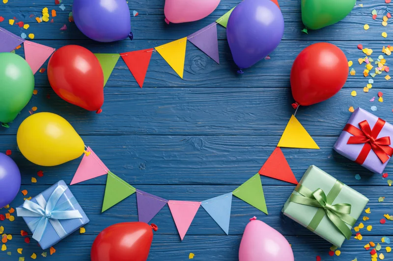 Colorful balloons and wrapped gifts arranged with bunting and confetti on blue wooden surface