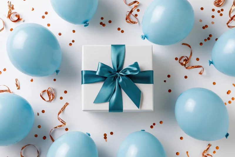 White gift box with blue ribbon surrounded by blue balloons and confetti on white surface