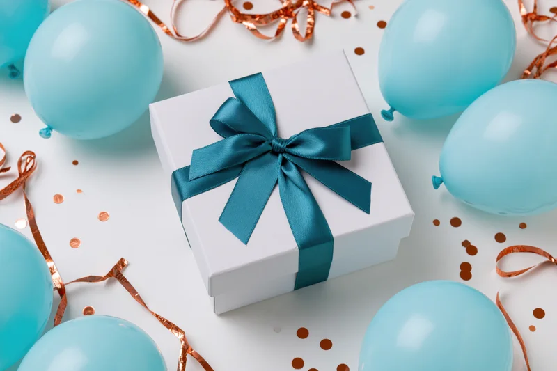 White gift box with teal ribbon and bow surrounded by blue balloons and copper streamers on white surface
