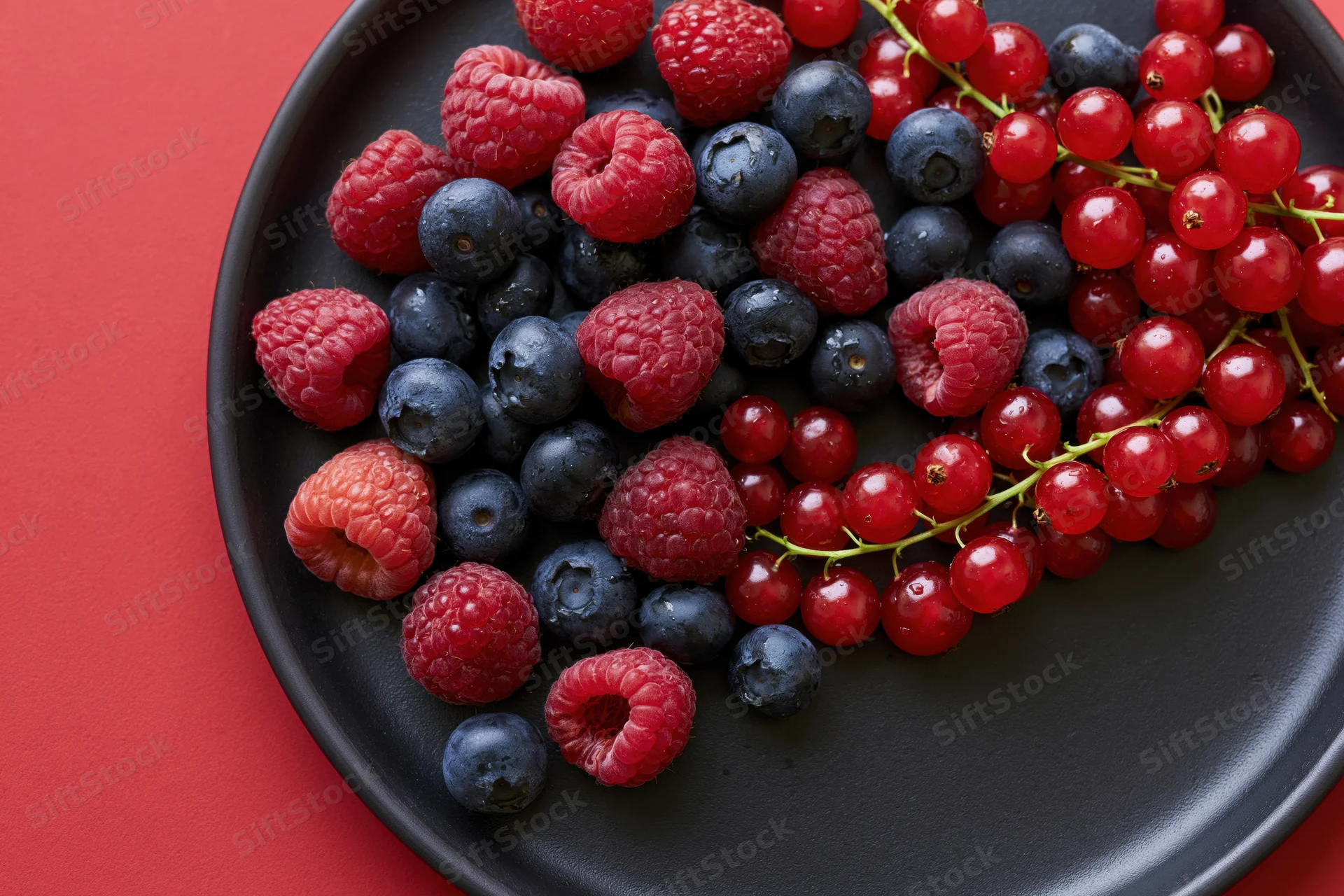 Fresh raspberries blueberries and redcurrants on a black ceramic plate against red background