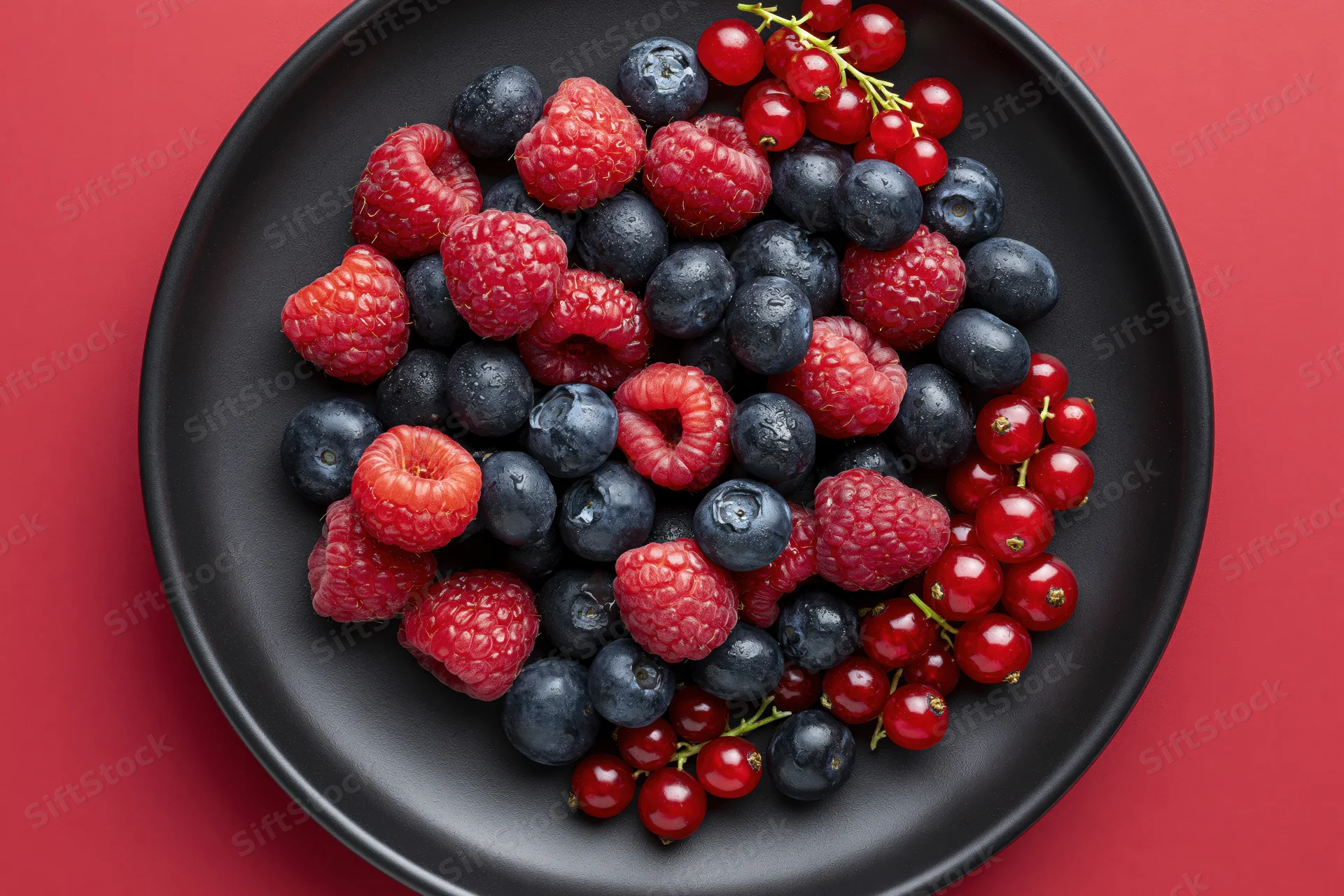 Fresh raspberries blueberries and redcurrants on a black ceramic plate viewed from above on a red surface