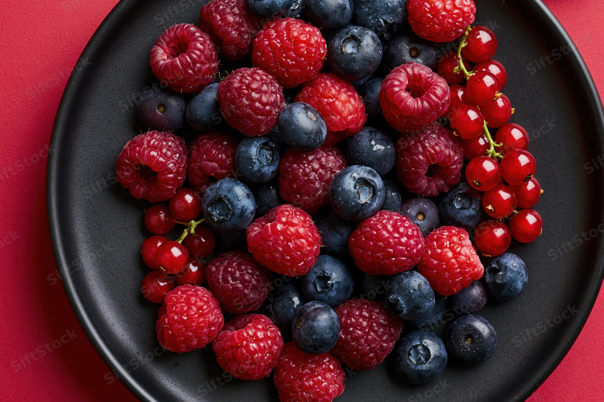 Fresh raspberries blueberries and redcurrants on a black ceramic plate against red background