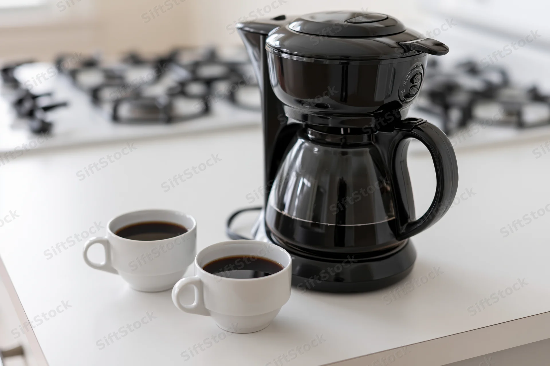 Black drip coffee maker with full glass carafe and two white coffee cups on kitchen countertop