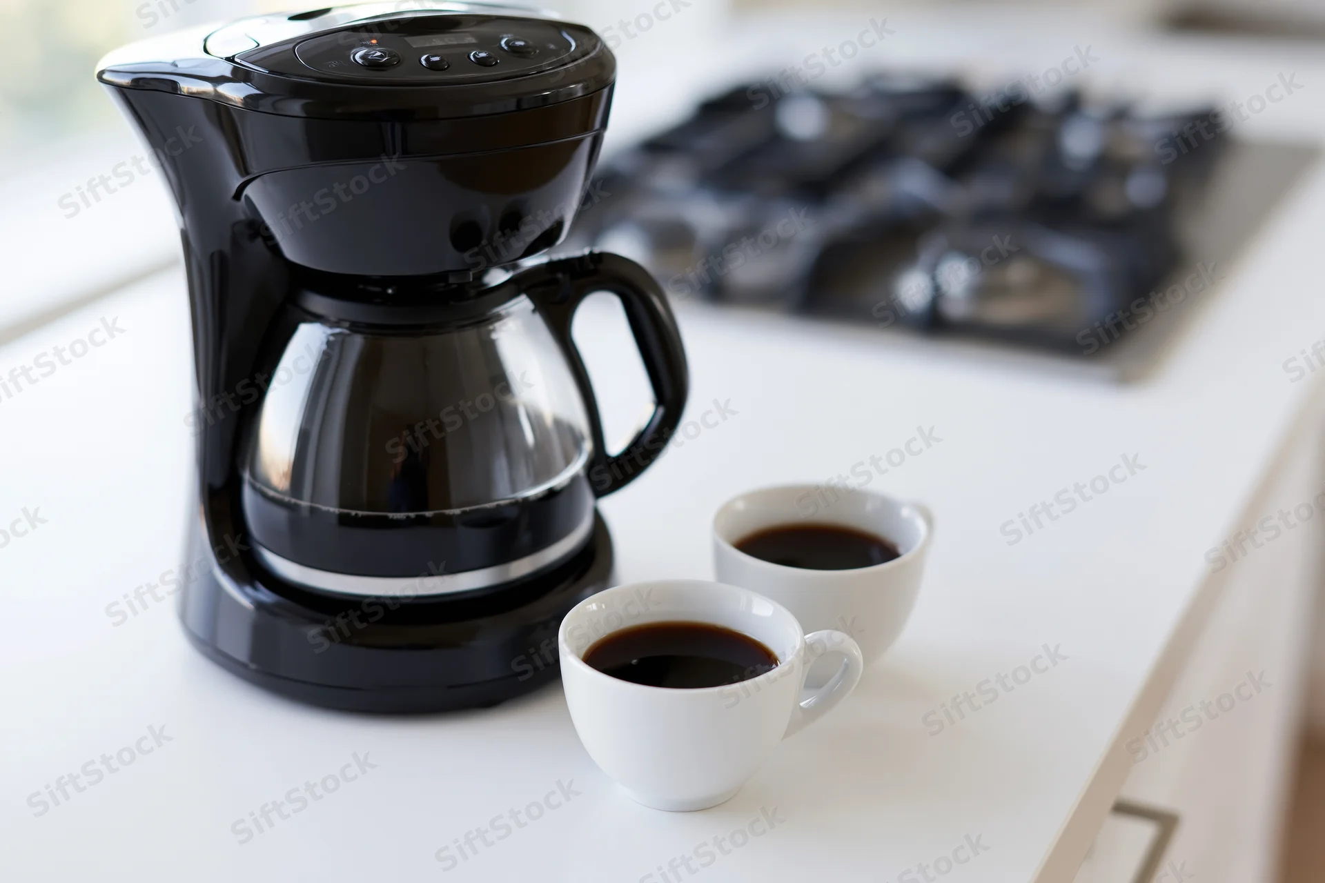 Black drip coffee maker with full glass carafe and two white coffee cups on white kitchen countertop