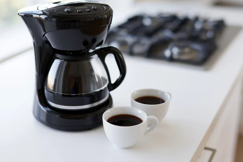 Black drip coffee maker with full glass carafe and two white coffee cups on white kitchen countertop