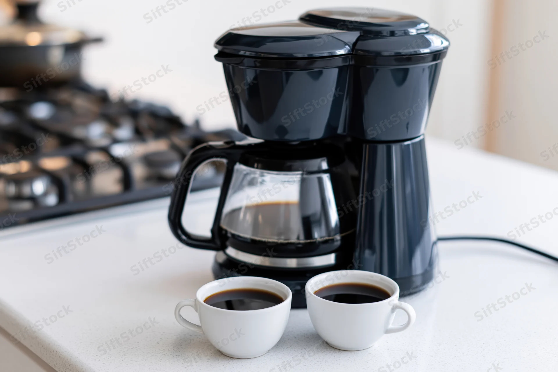 Black drip coffee maker with full glass carafe and two white cups of coffee on kitchen countertop near gas stove