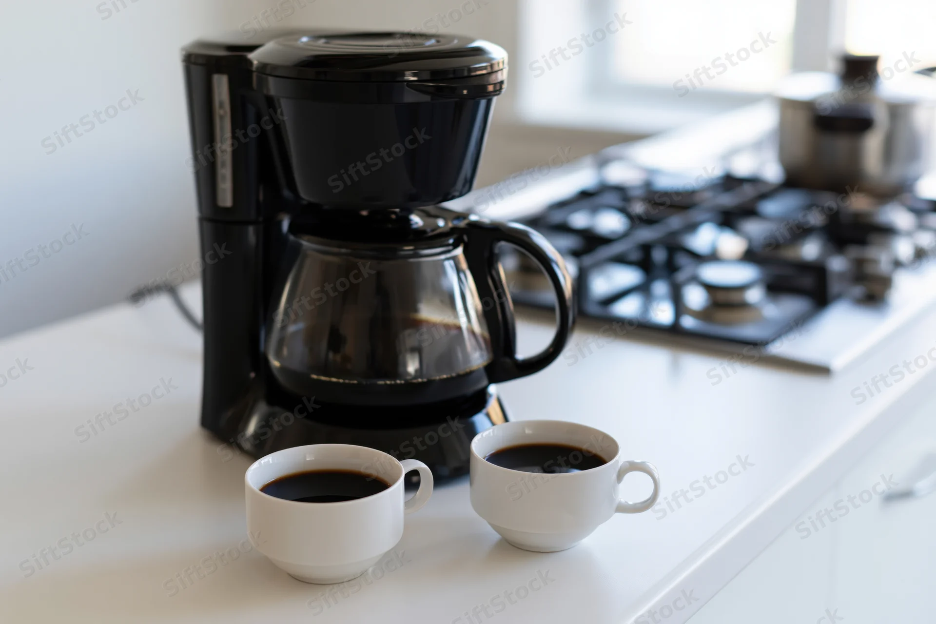 Black drip coffee maker with full glass carafe and two white cups of coffee on kitchen countertop near gas stove