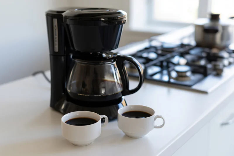 Black drip coffee maker with full glass carafe and two white cups of coffee on kitchen countertop near gas stove