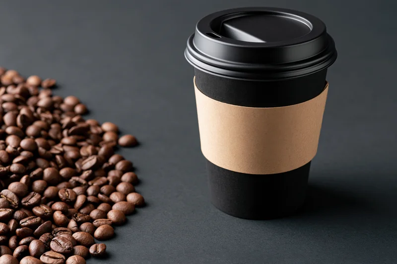 Black paper takeaway coffee cup with kraft sleeve beside roasted coffee beans on dark surface
