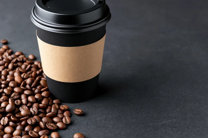 Black paper takeaway coffee cup with blank kraft sleeve and scattered coffee beans on dark surface