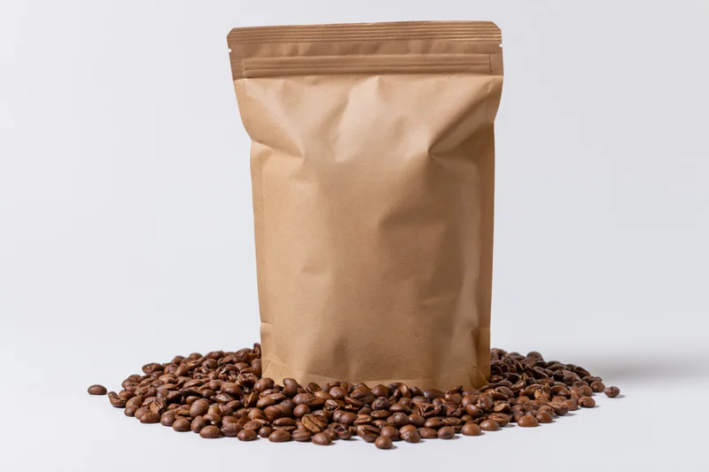 Brown kraft paper coffee bag standing upright with roasted coffee beans scattered around on white background