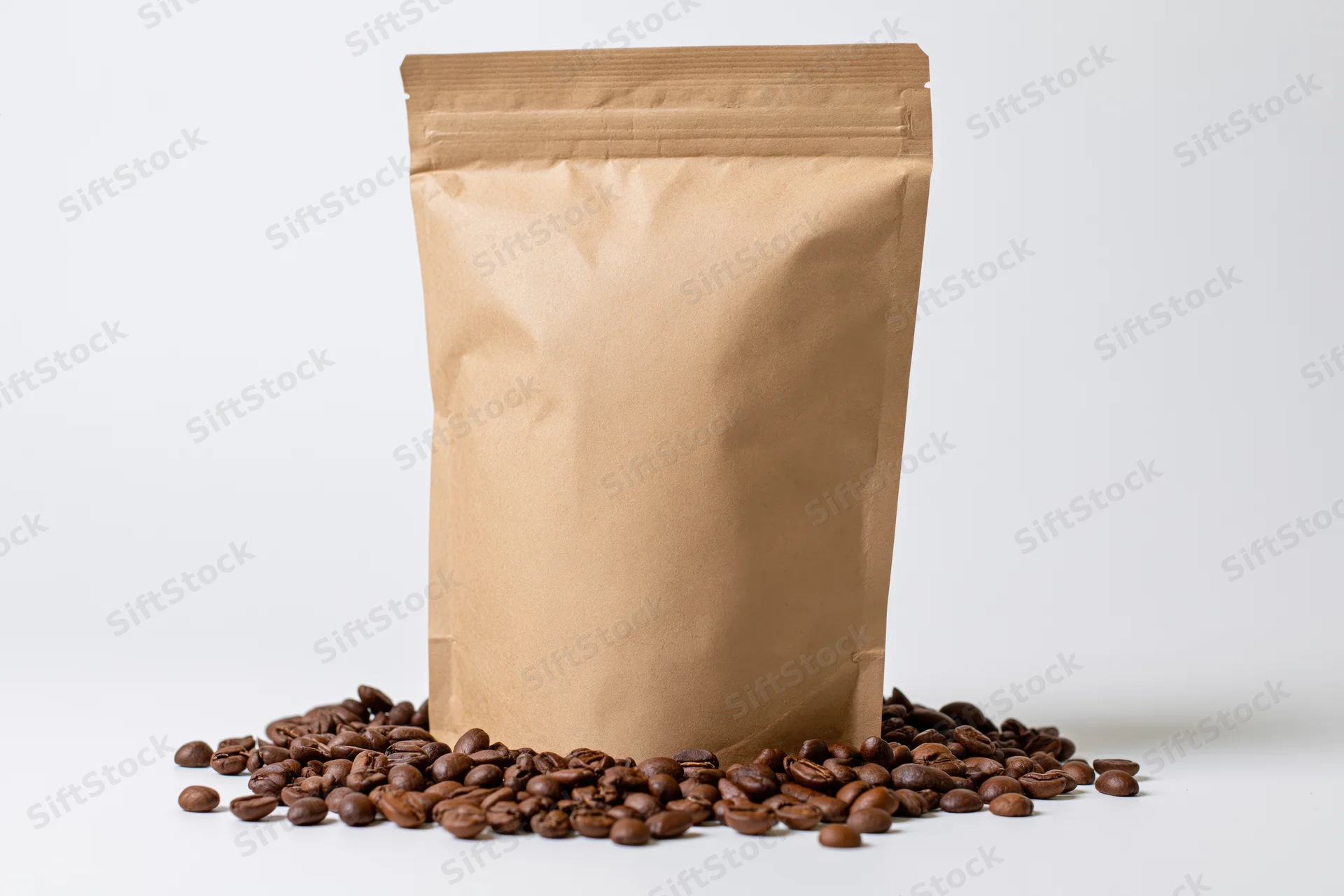 Blank kraft paper coffee bag standing upright with scattered coffee beans on white surface
