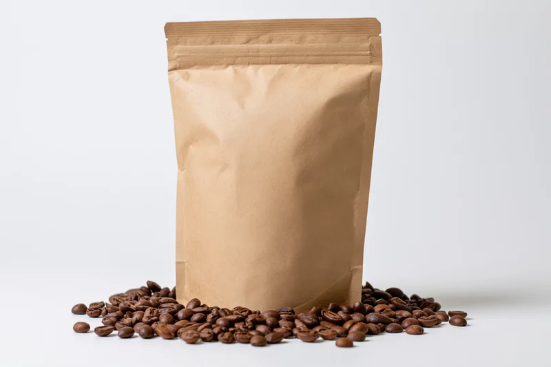 Blank kraft paper coffee bag standing upright with scattered coffee beans on white surface