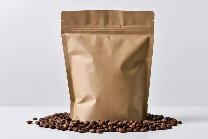 Blank kraft paper coffee bag standing upright with scattered coffee beans on white surface