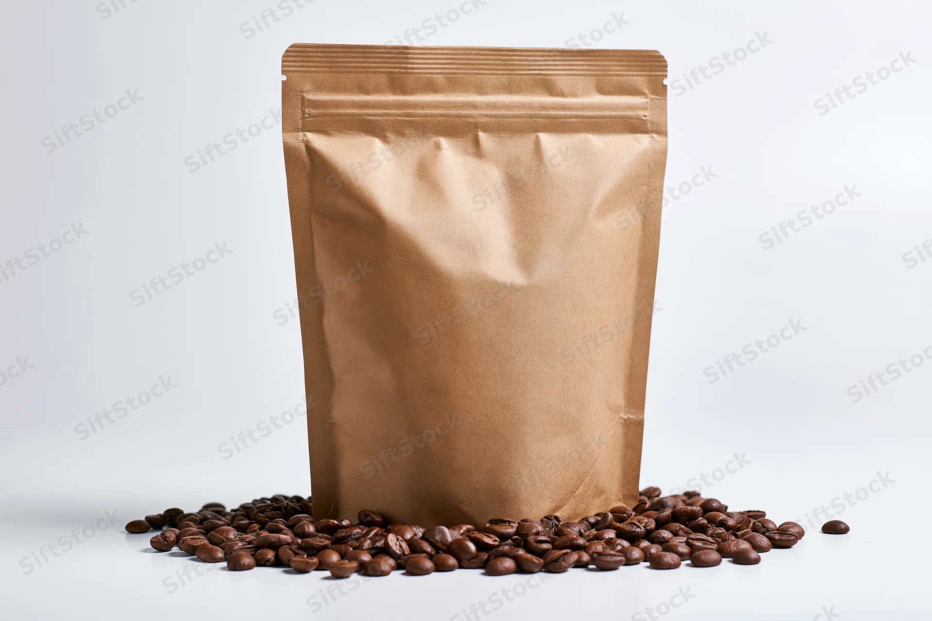 Blank kraft paper coffee bag standing upright with scattered roasted beans on white surface