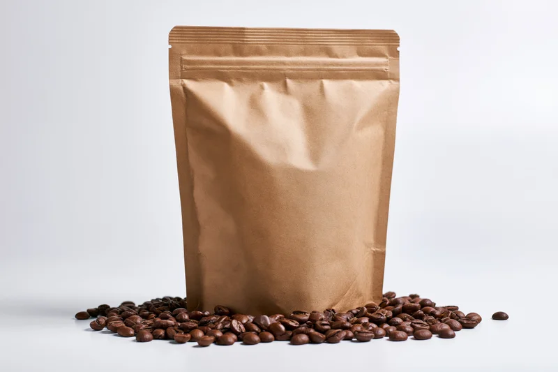 Blank kraft paper coffee bag standing upright with scattered roasted beans on white surface