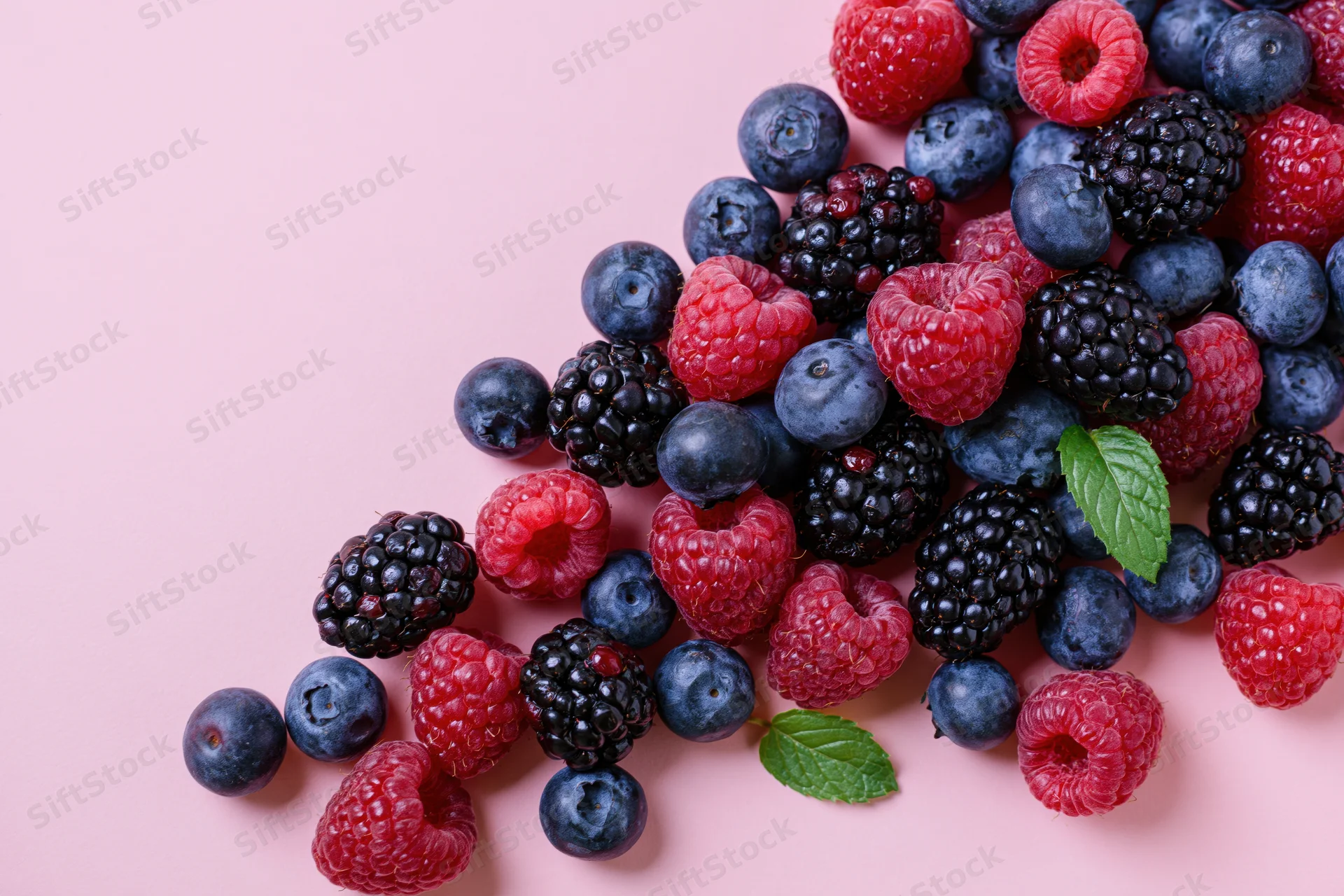 Blueberries, blackberries and raspberries scattered on a pink surface with mint leaves