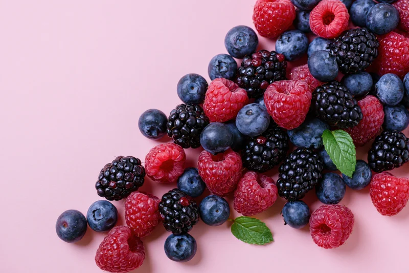 Blueberries, blackberries and raspberries scattered on a pink surface with mint leaves