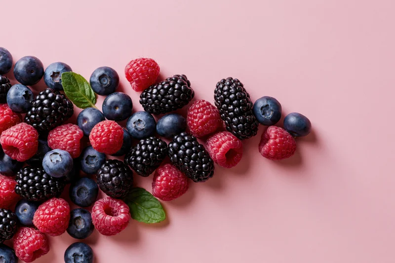 Blueberries, blackberries and raspberries scattered on a pink surface with mint leaves and copyspace