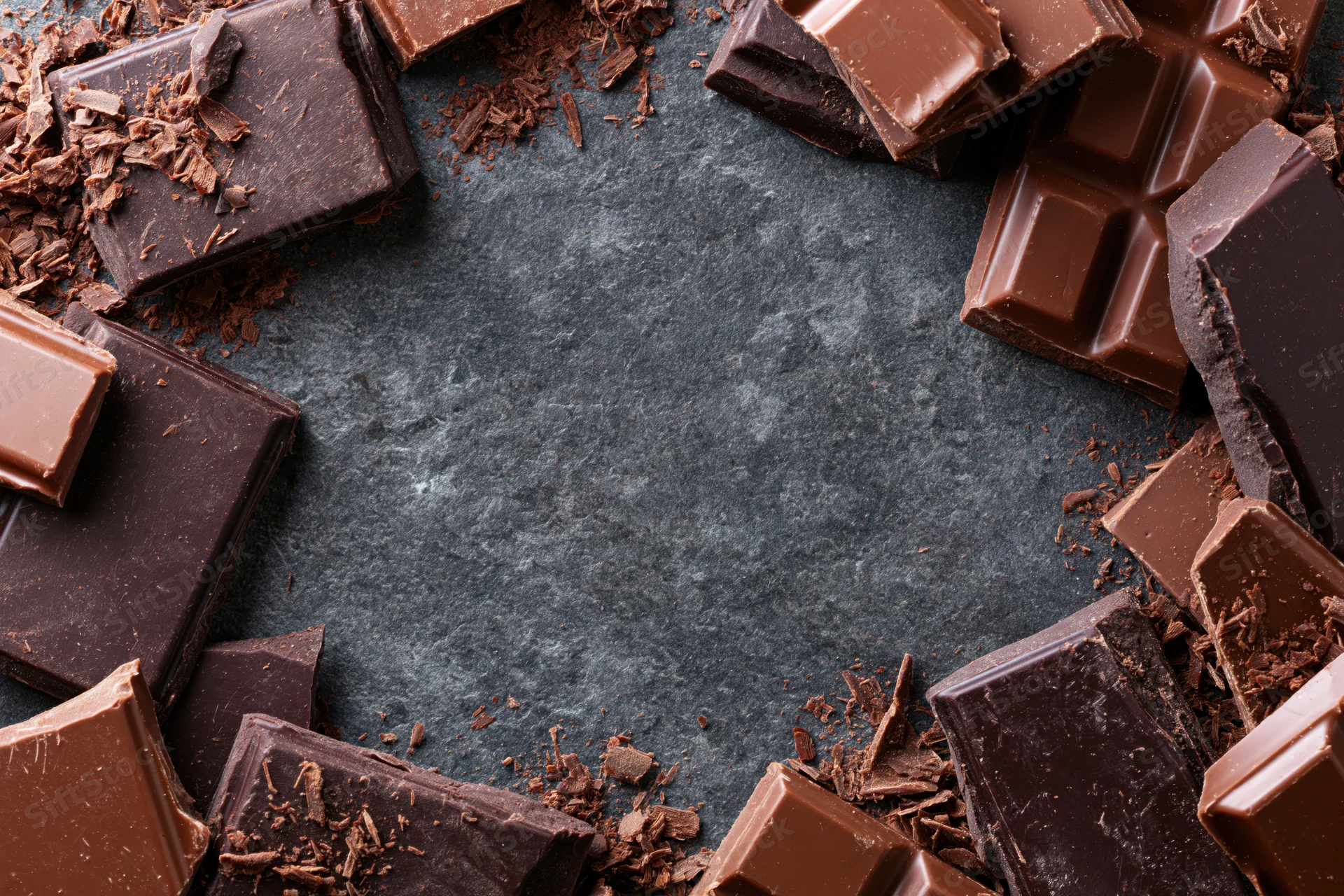 Broken dark and milk chocolate bar pieces scattered around a dark surface forming a frame with copyspace in center