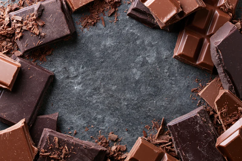 Broken dark and milk chocolate bar pieces scattered around a dark surface forming a frame with copyspace in center