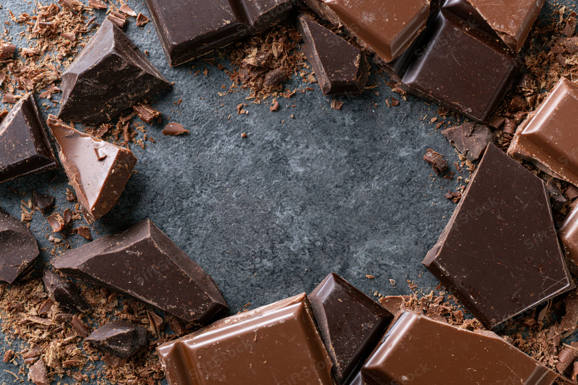 Broken dark and milk chocolate pieces scattered in a loose frame around a central copyspace on a dark stone surface
