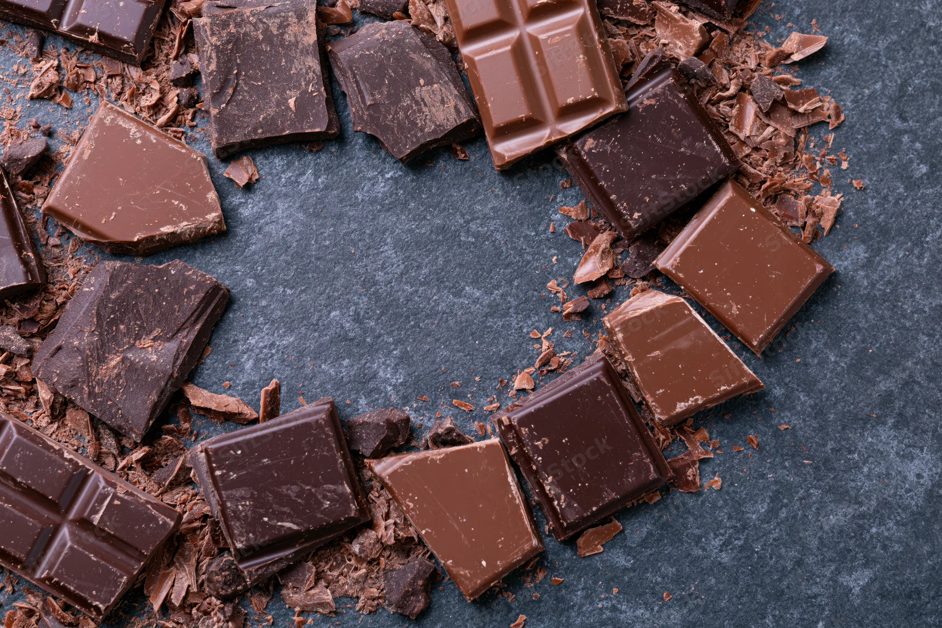 Broken dark and milk chocolate pieces scattered in a loose circle on a gray stone surface with central copyspace