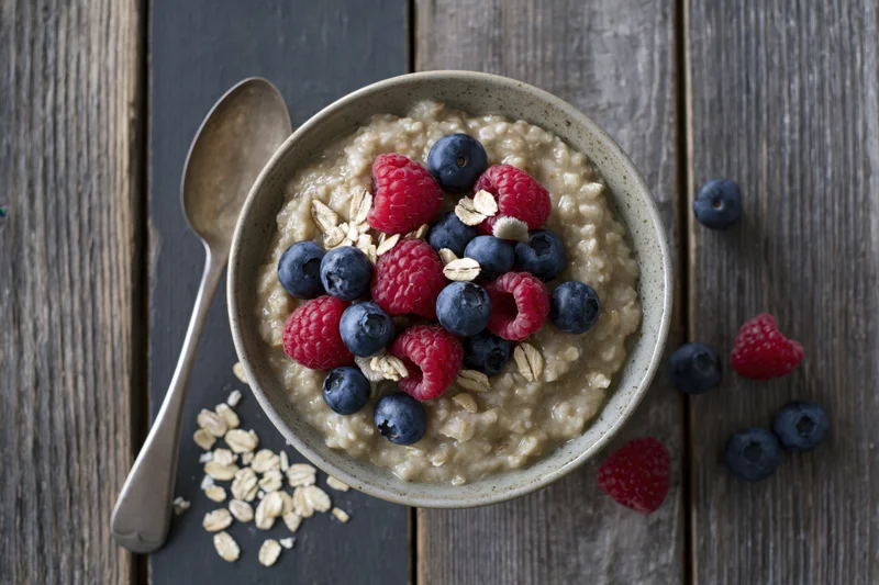 Ceramic bowl of oatmeal porridge topped with fresh raspberries and blueberries on wooden table with spoon and scattered oats